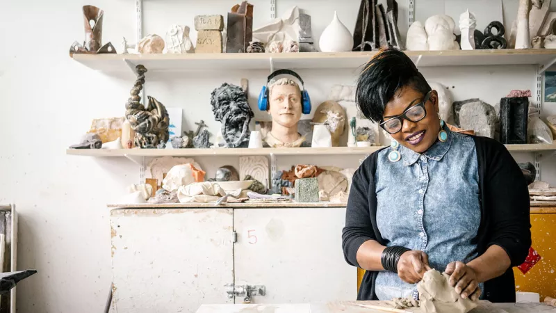 Poster of Female student standing to the right of the image, in a sculpture workshop, using some clay